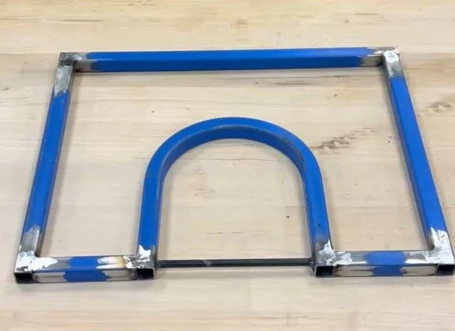 A welded steel pressing frame on a workbench: a rectangle with an arc bridging its base, the paint sandblasted back to bright metal at every weld.