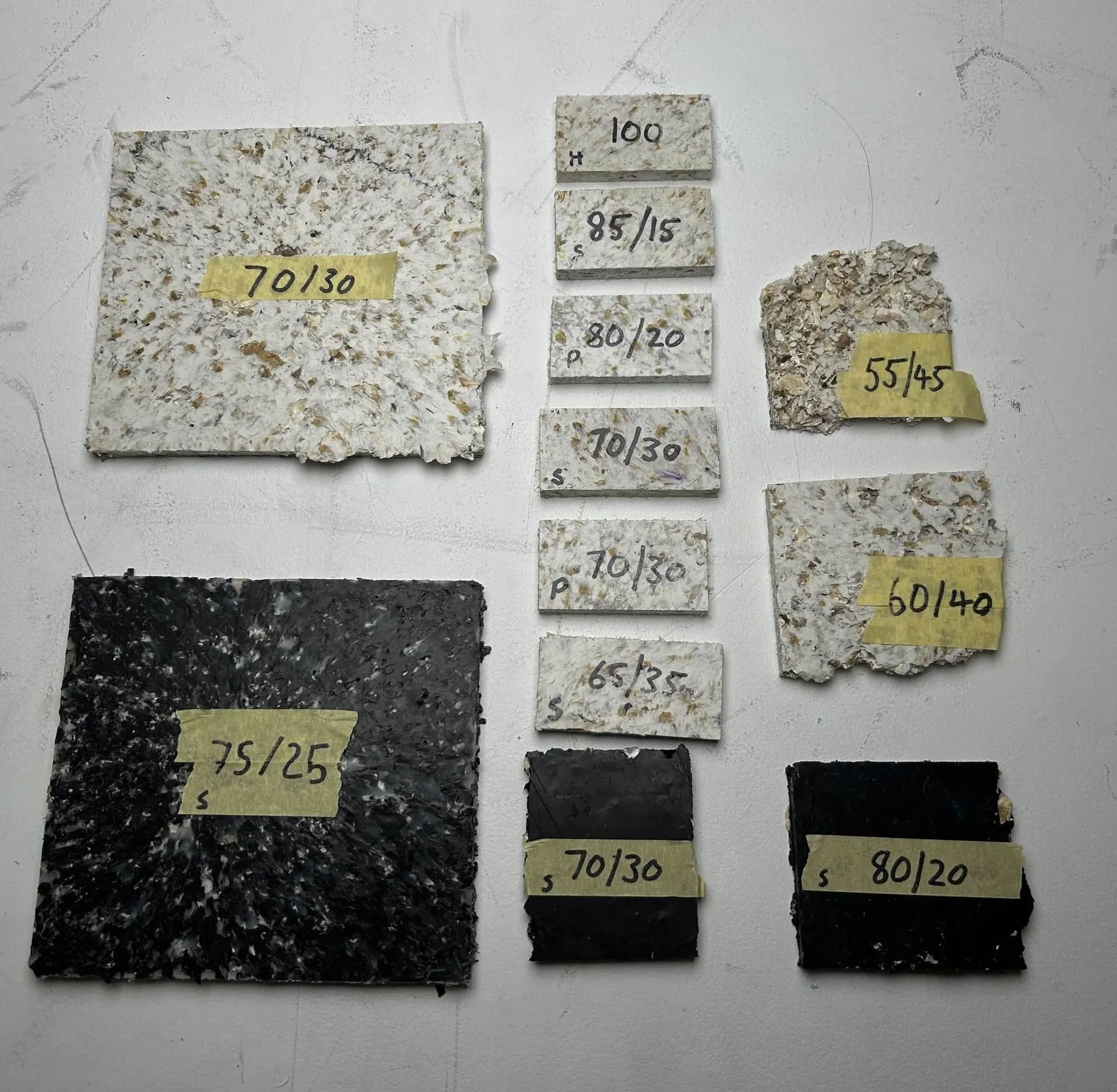 Twelve composite test tiles laid out on a concrete floor, each labelled in marker with its HDPE-to-shell ratio: 100, 85/15, 80/20, 75/25, 70/30, 65/35, 60/40, 55/45.
